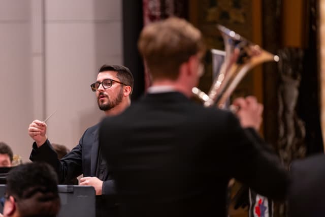 Conductor with baton leads band, framed by a blurred brass player in the foreground