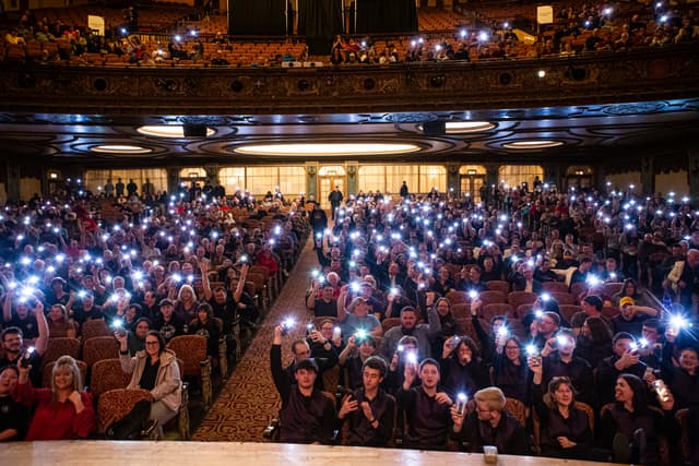 Audience in an ornate theatre holding up lit phones toward the stage