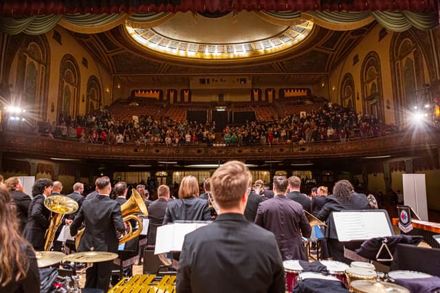 Band seen from behind on stage, facing applauding audience in ornate historic theatre balcony