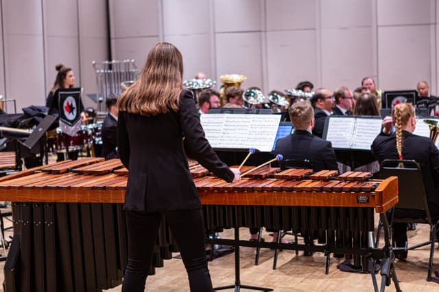 Marimba player seen from behind with brass band musicians seated on stage