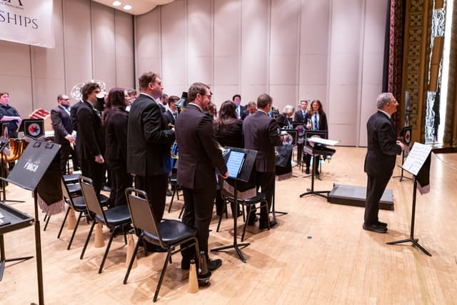 Brass band members standing on a concert stage with instruments, music stands and a conductor
