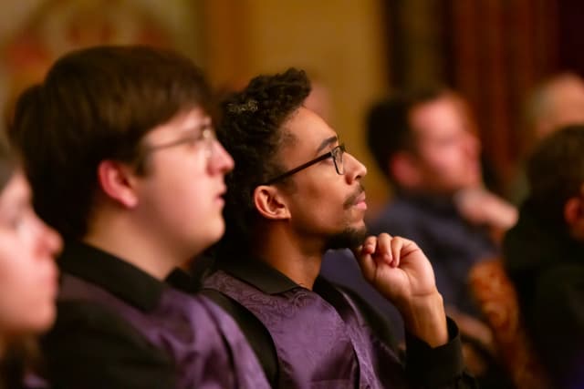 Attentive audience members in purple vests watching a performance in a warmly lit hall