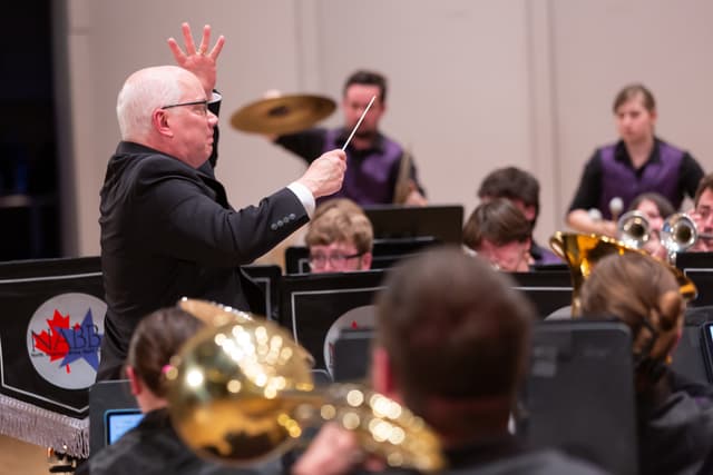 Conductor with baton leads a brass band as a cymbal player and brass musicians perform.