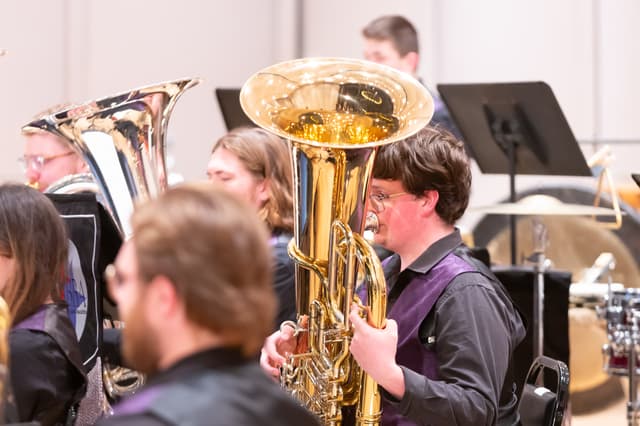 Tuba player performing with brass band members, music stands and percussion behind on stage