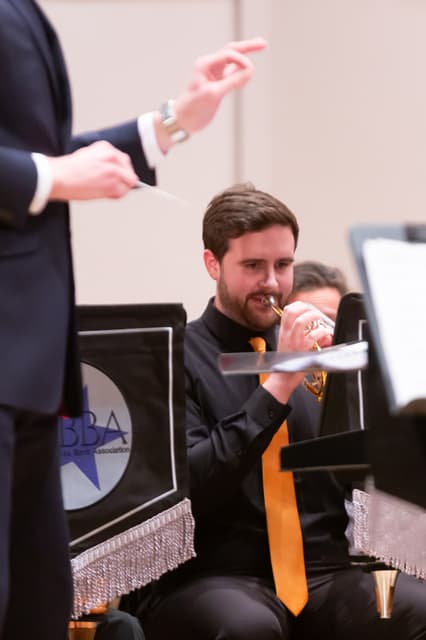 Cornet player performing on stage as a conductor's hands direct the band.
