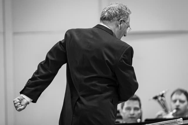 Conductor from behind with clenched fist leading brass band players in black and white