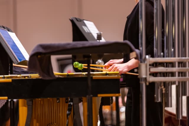 Percussionist's hands holding green mallets over a vibraphone beside tubular bells and music stands