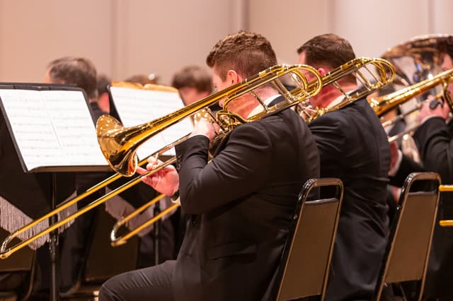 Trombone players performing in a brass band, seen from behind, with sheet music on stands