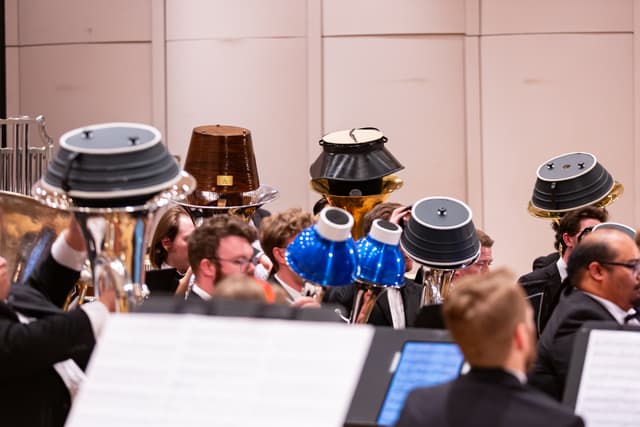 Brass band tubas with mutes on their bells, players seated in front of music stands