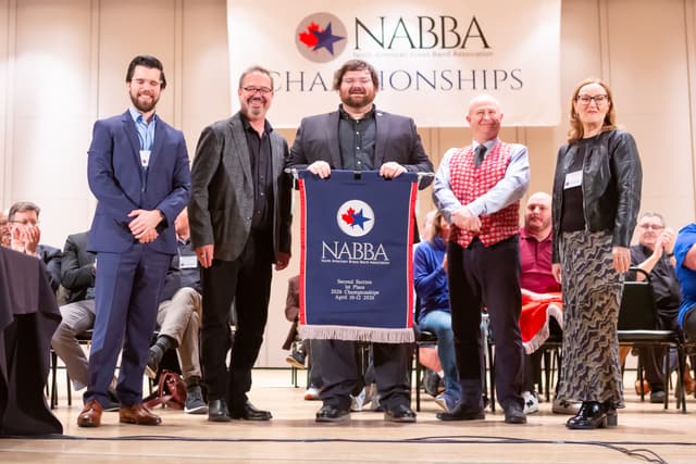 Five people on stage holding a NABBA Second Section 1st Place banner as audience applauds