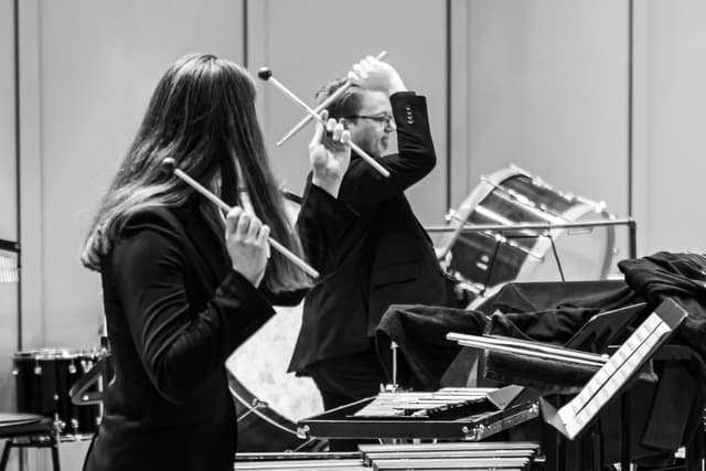 Two percussionists raise mallets while playing on stage near keyboard percussion and drums