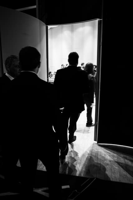 Silhouetted people in suits walking from a dark backstage through a doorway into a bright hall