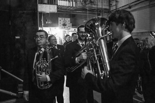 Brass players holding euphoniums and cornet walk through a backstage corridor in black and white.