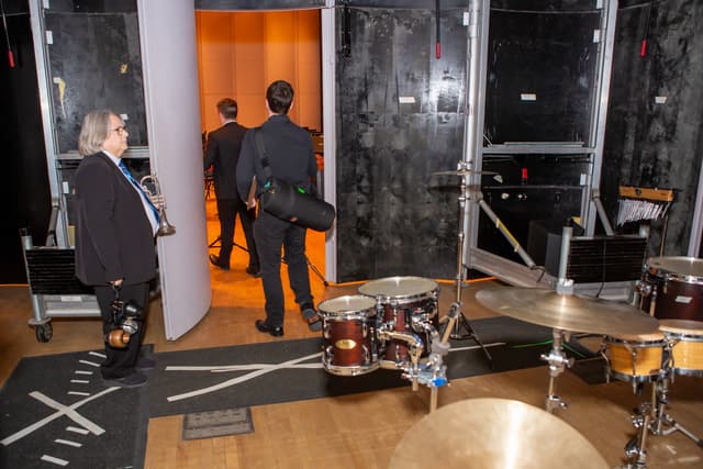 Musicians walking through a backstage door toward the stage, with a drum kit in the foreground