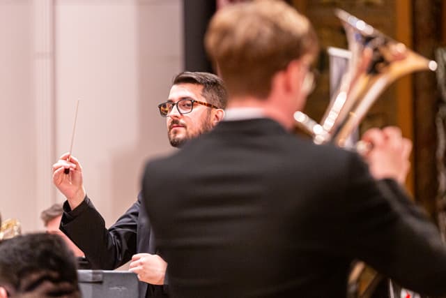 Conductor with raised baton glances past a blurred tuba player during a brass band performance.