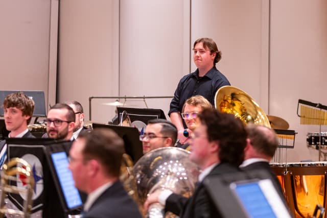 Brass band players seated on stage with tubas and timpani while one member stands behind them