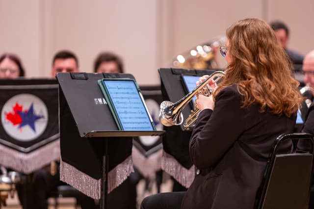 Cornet player reading a tablet on a music stand, with band members and banners behind
