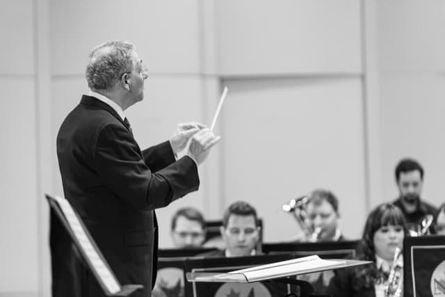 Conductor with raised baton leading seated brass band musicians in black and white