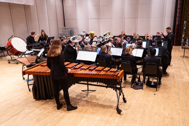 Brass band performing on stage with conductor, marimba in foreground and tubas and percussion behind