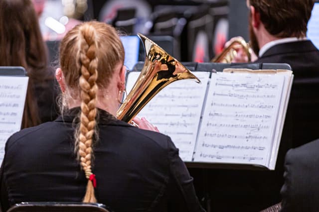 Back view of a brass musician with braided hair playing from sheet music on a stand