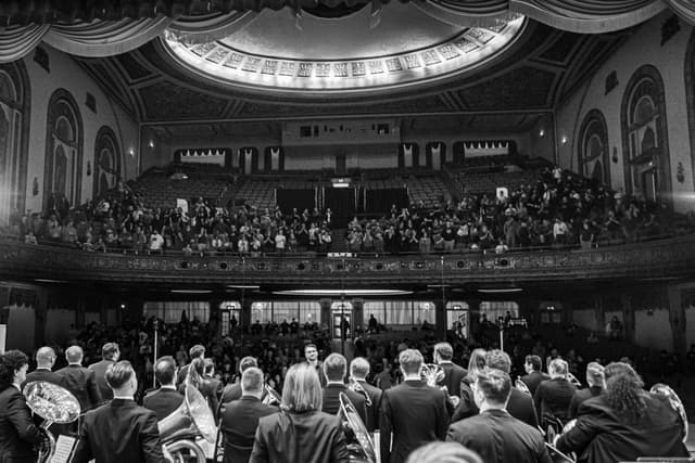 Brass band seen from behind on stage facing a standing audience in an ornate theatre