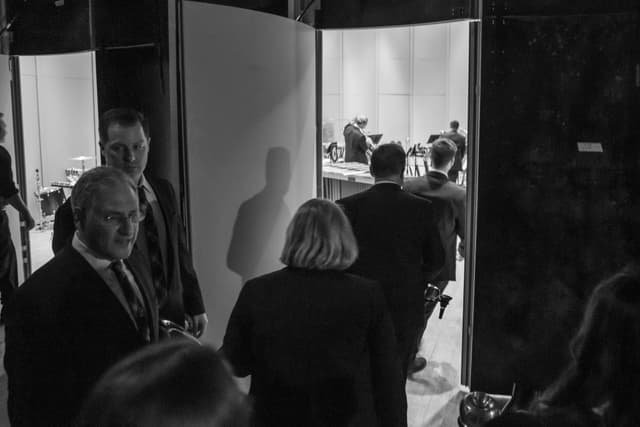 Musicians in black attire walk backstage through a doorway toward a stage with percussion