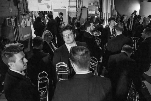 Black and white photo of brass players in suits crowded backstage with trombones and horns
