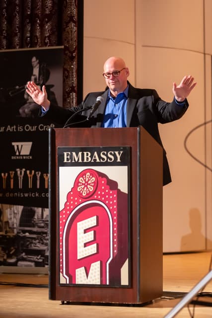 Speaker with raised arms at an Embassy podium beside a Denis Wick banner on stage