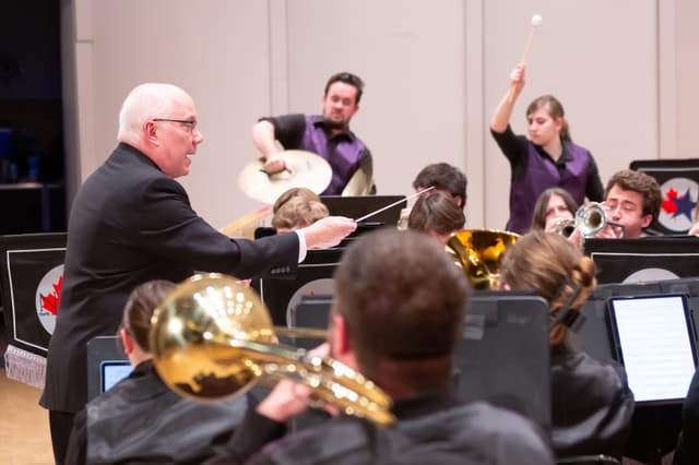 Conductor directing brass band while percussionists play cymbals and mallet, trombone in foreground