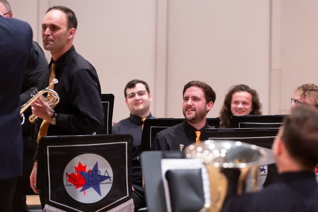 Smiling brass band members seated on stage while a standing cornet player holds his instrument.
