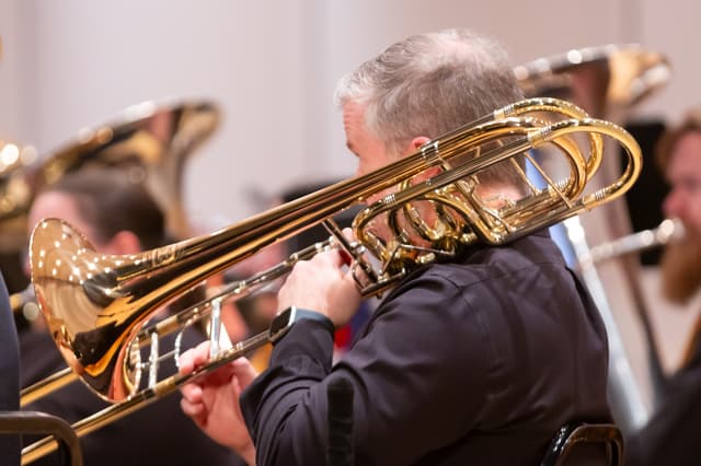 Trombonist playing a gleaming bass trombone with tubas blurred in the background