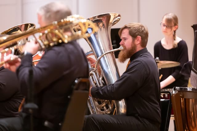 Tuba player performing beside trombones, with a percussionist in the background on stage