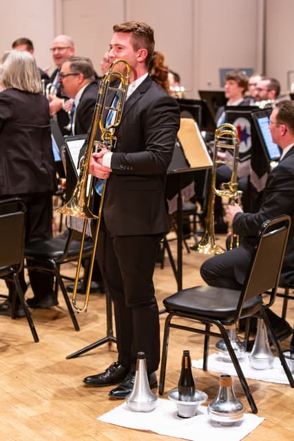 A standing trombonist holds his instrument beside a row of mutes on a cloth, with band members behind.