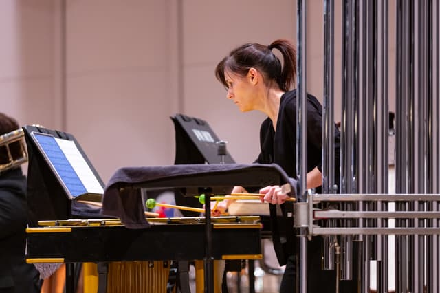 Percussionist playing vibraphone with mallets next to tubular bells, reading music on a stand