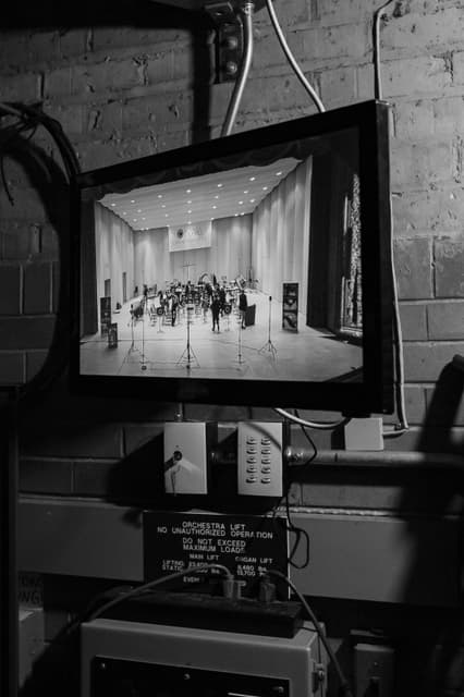Backstage monitor showing the NABBA Championships stage, with an orchestra lift warning sign below