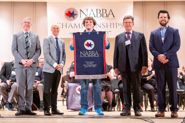 Five people on stage holding a NABBA Third Section 1st Place banner at the championships awards ceremony.