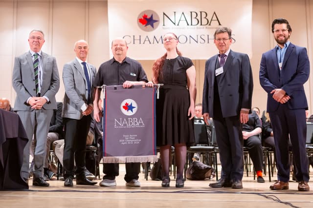 Six people on stage holding a NABBA third place banner at the championships awards ceremony