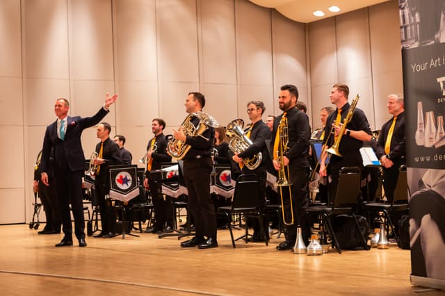 Conductor gesturing to a standing brass band on stage during applause