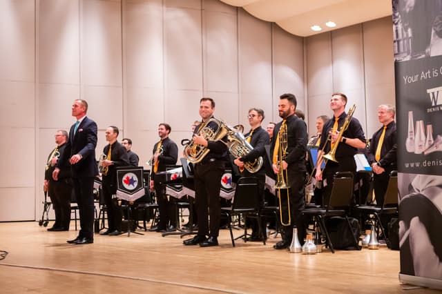 Brass band members standing on stage with instruments, smiling, beside their conductor after a performance.