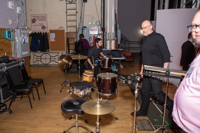 Percussion equipment and musicians setting up in a backstage area with brick walls