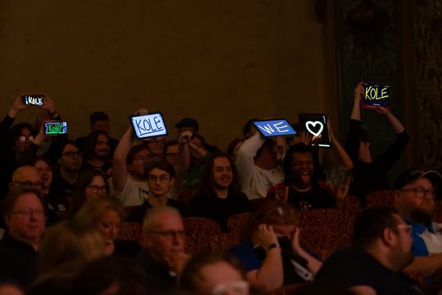 Audience members raise glowing tablets and phones showing "KOLE", "WE" and a heart in a dark theatre.