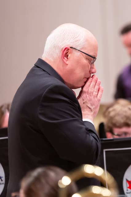 Man in dark suit with hands clasped near his face among seated brass band players