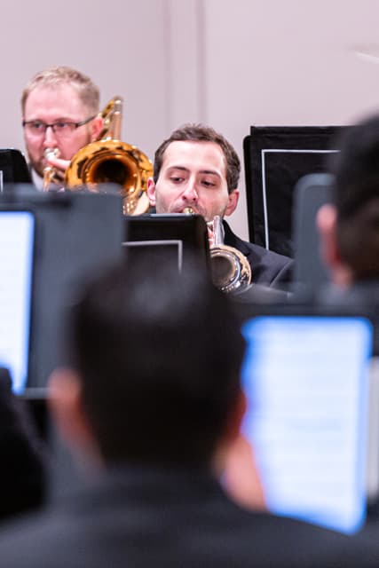 Trumpet and trombone players performing between music stands, with blurred band members in the foreground