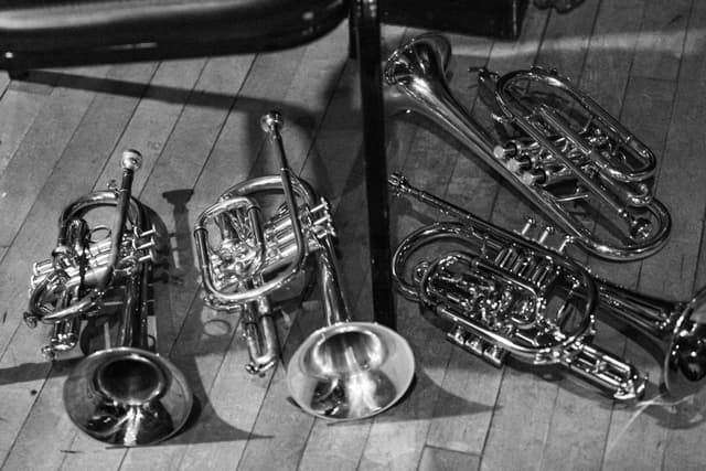 Silver cornets and trumpets lying on a wooden floor beside a black stand, in black and white