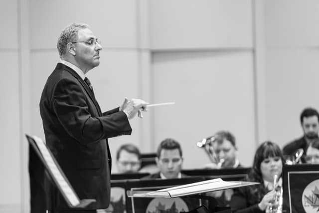 Conductor with baton leads a brass band seated behind music stands in black and white.