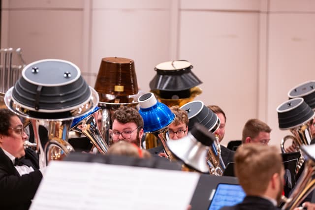Brass band players with mutes over tuba and euphonium bells, reading music on stage