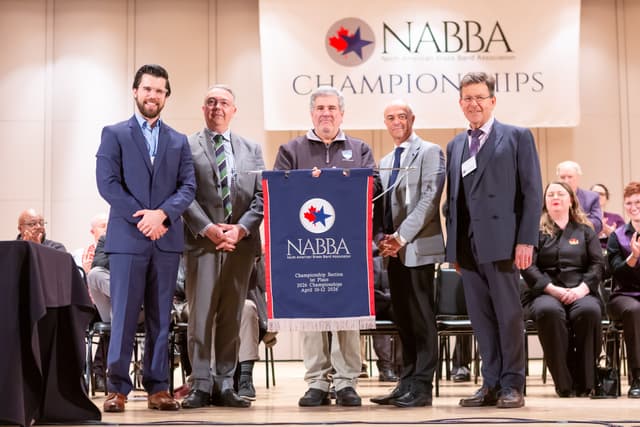 Five men on stage holding a NABBA first place championship banner while others applaud behind them