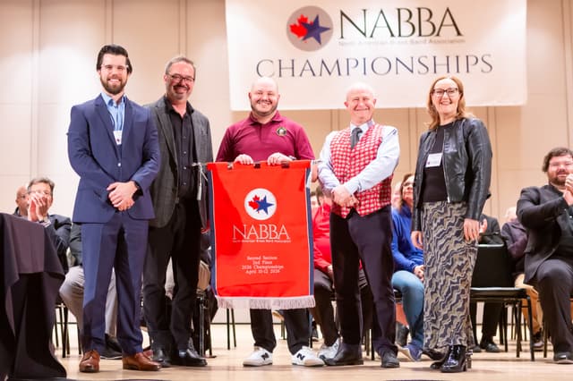 Five people on stage holding a red NABBA second place banner while seated audience applauds