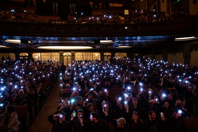 Theatre audience raising glowing phone lights in a dark ornate auditorium