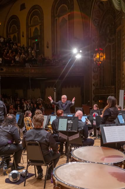 Conductor with open arms facing applauding audience, brass band seated on an ornate theatre stage